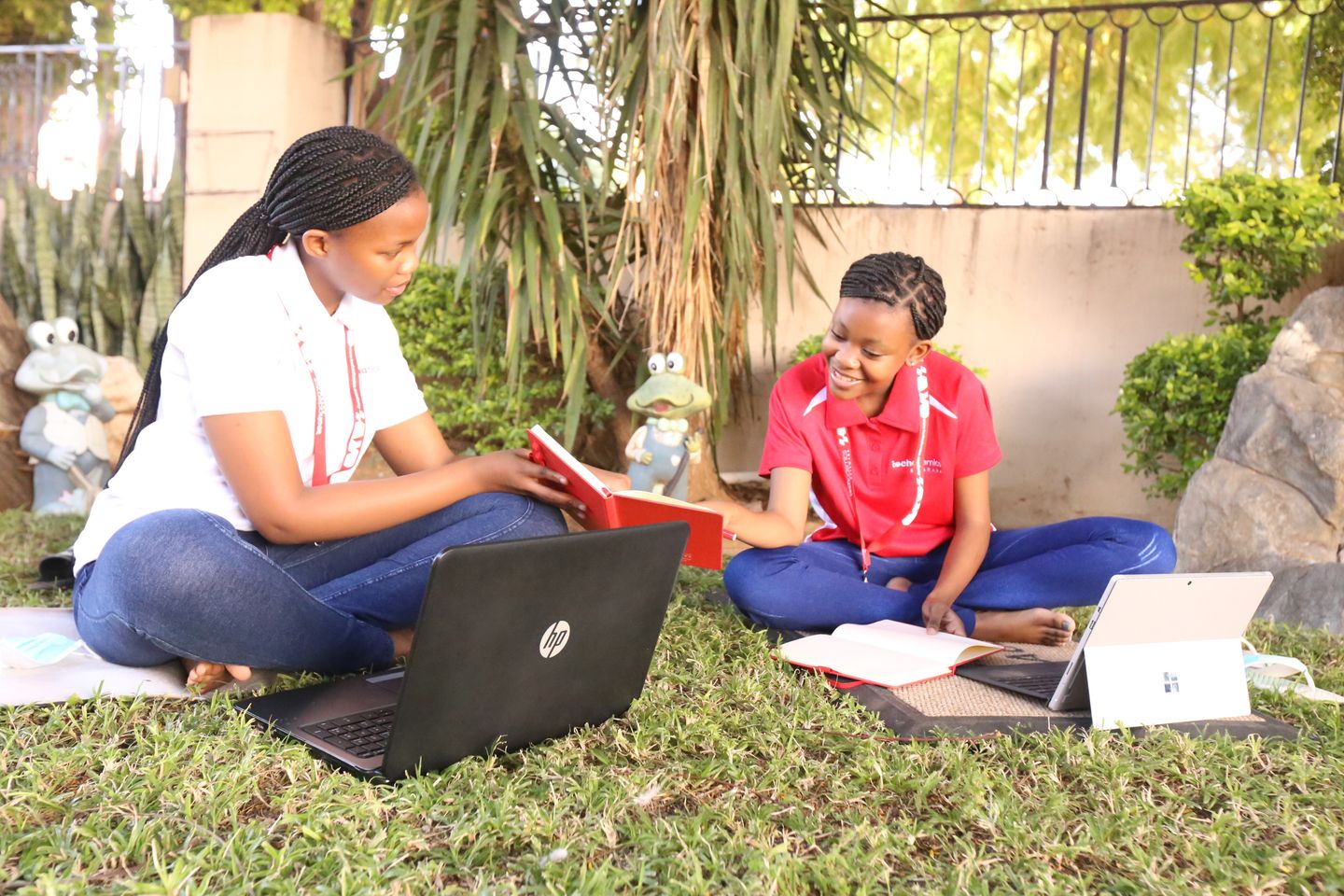 Two students in Techademics Academy uniforms sitting on the grass reviewing a notebook together, laptops open beside them